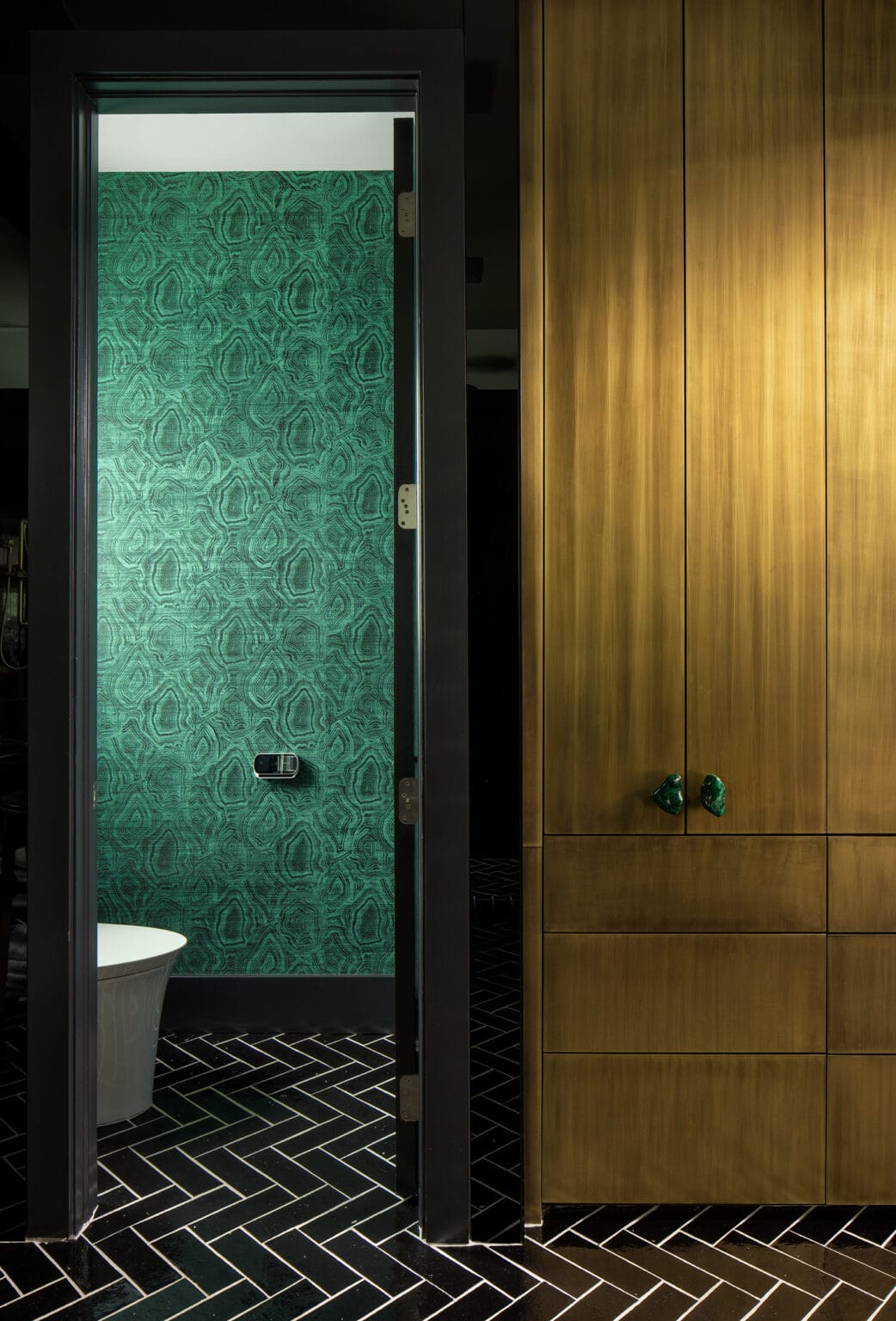 Brass-finished cabinetry beside a doorway to a bathroom with green marbled wallpaper and a freestanding tub