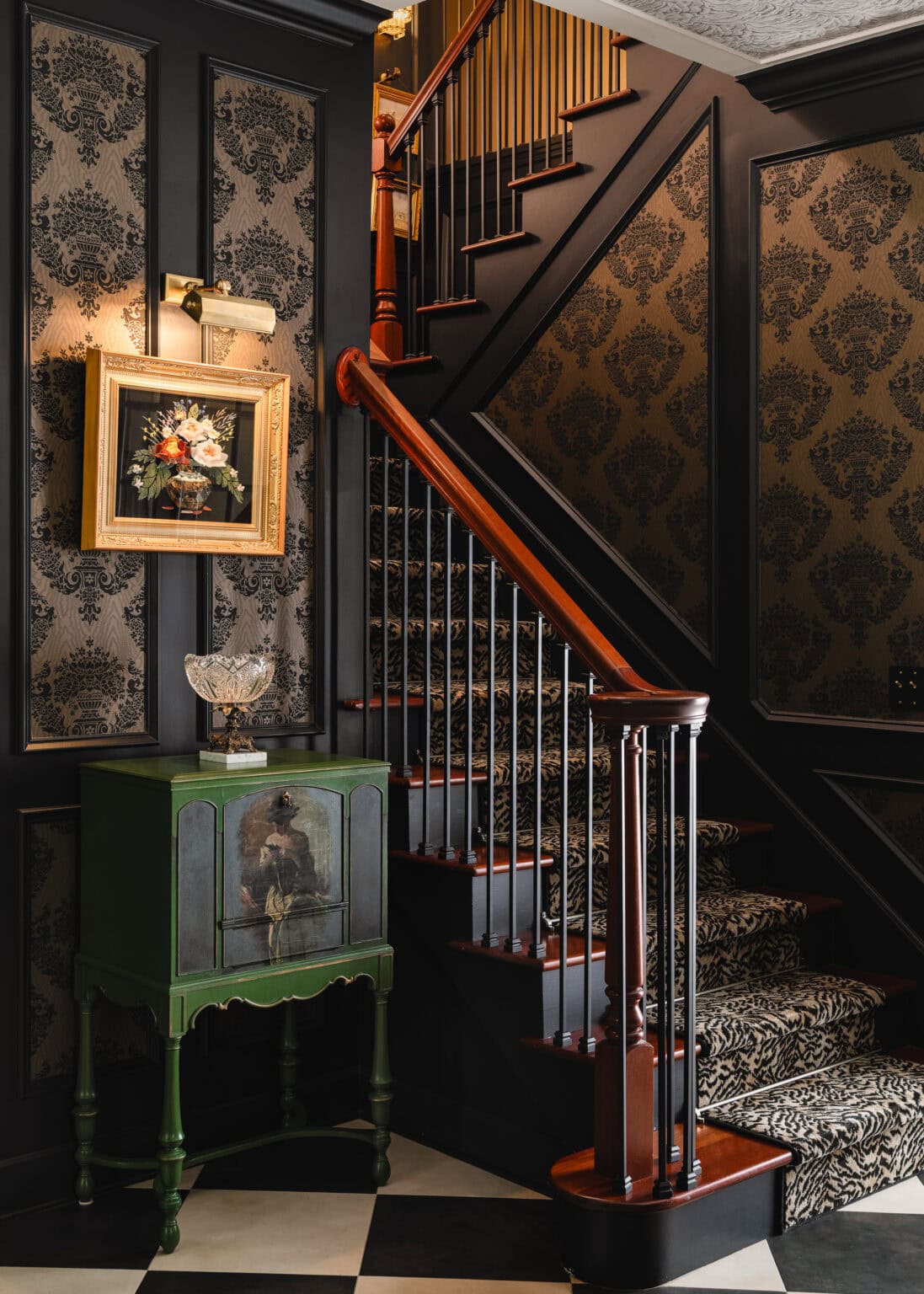Dark damask-papered stair hall with a red-railed staircase, a green painted chest and a gilt-framed painting