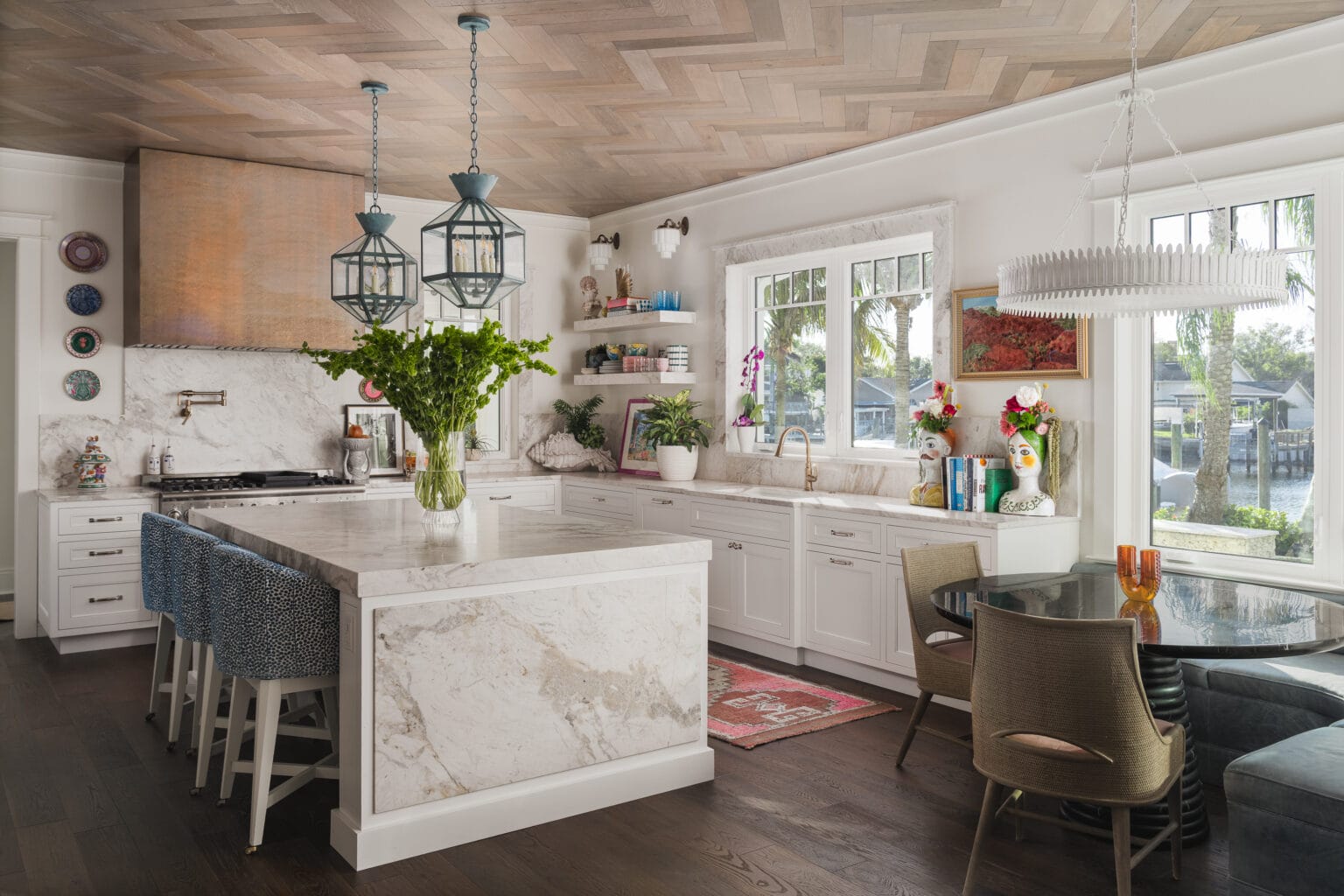 White kitchen with a marble waterfall island, lantern pendants, blue counter stools and a breakfast table by the windows