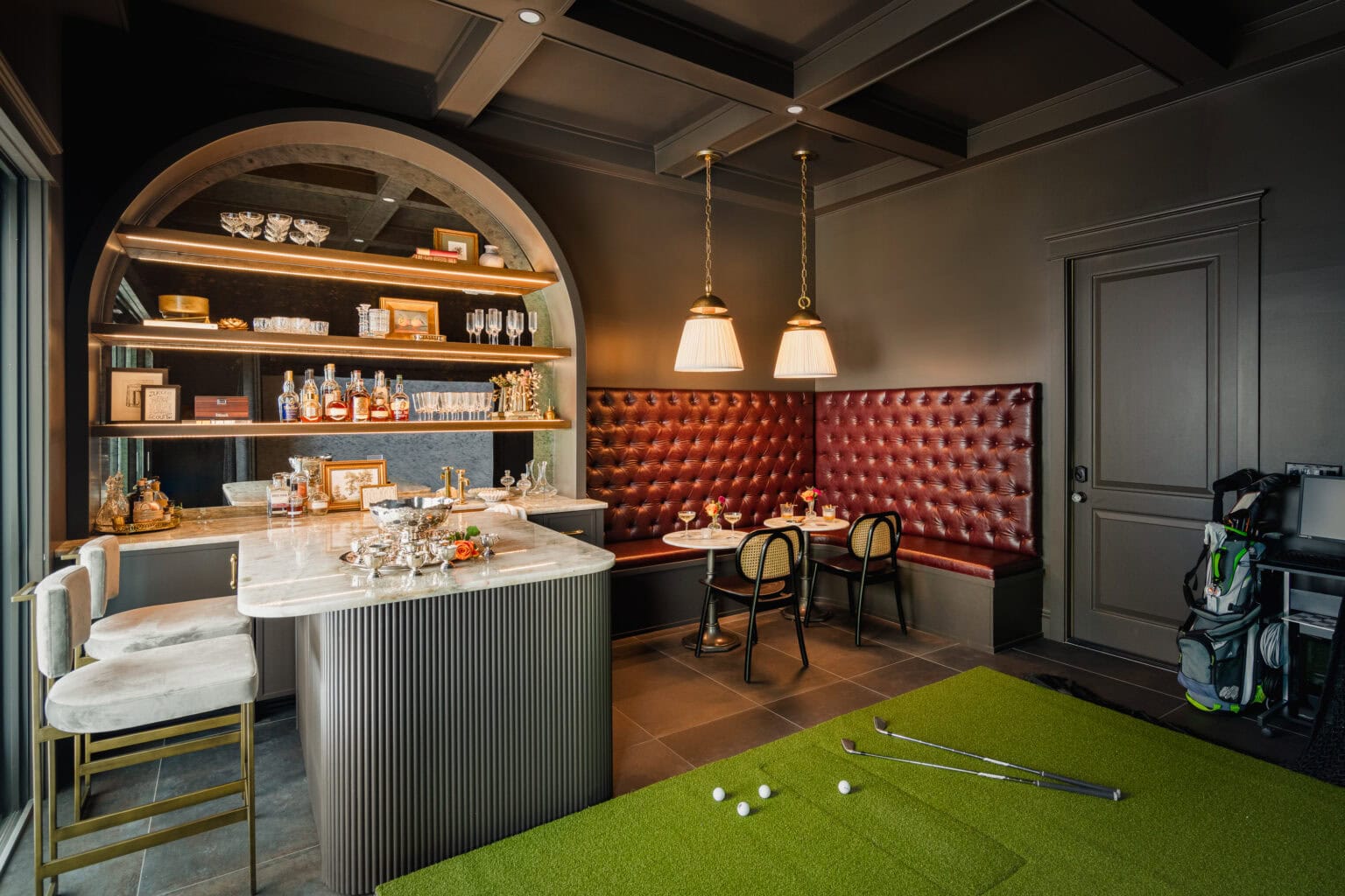 Wide view of a golf lounge with an arched bar, oxblood tufted banquette, coffered ceiling and indoor putting green