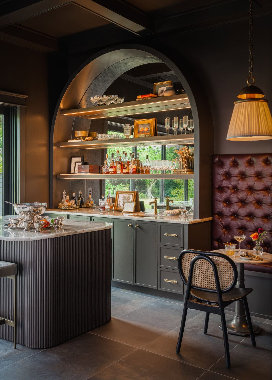 Home bar with an arched, lit shelving niche, marble counter and oxblood tufted banquette