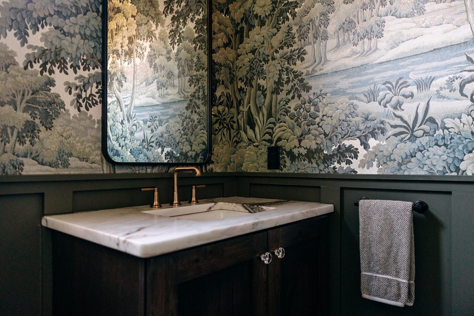 Powder room with scenic green wallpaper, dark green wainscoting and a marble vanity with a brass faucet
