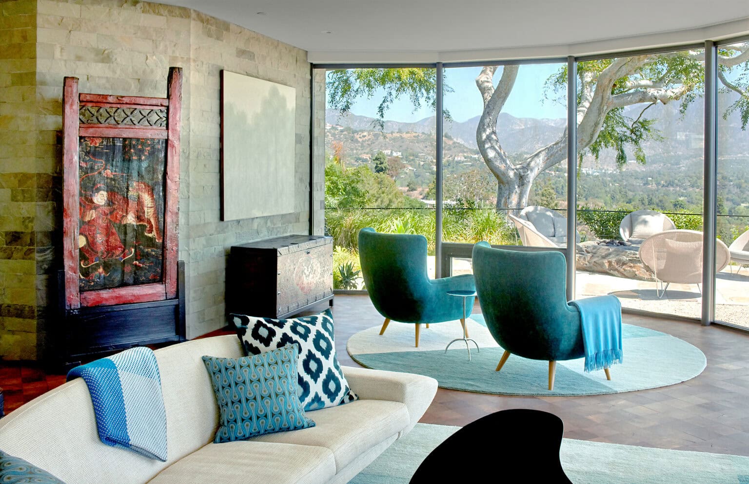 Curved glass living room with mountain views, a white sofa, teal lounge chairs and a stone accent wall