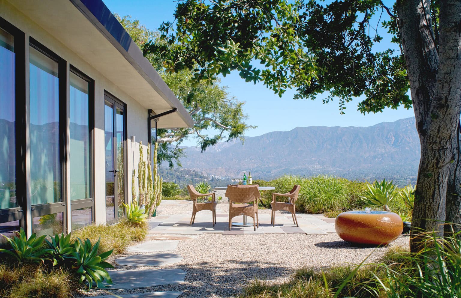 Stone terrace beside a modern glass home with outdoor seating, an oak tree and mountain views