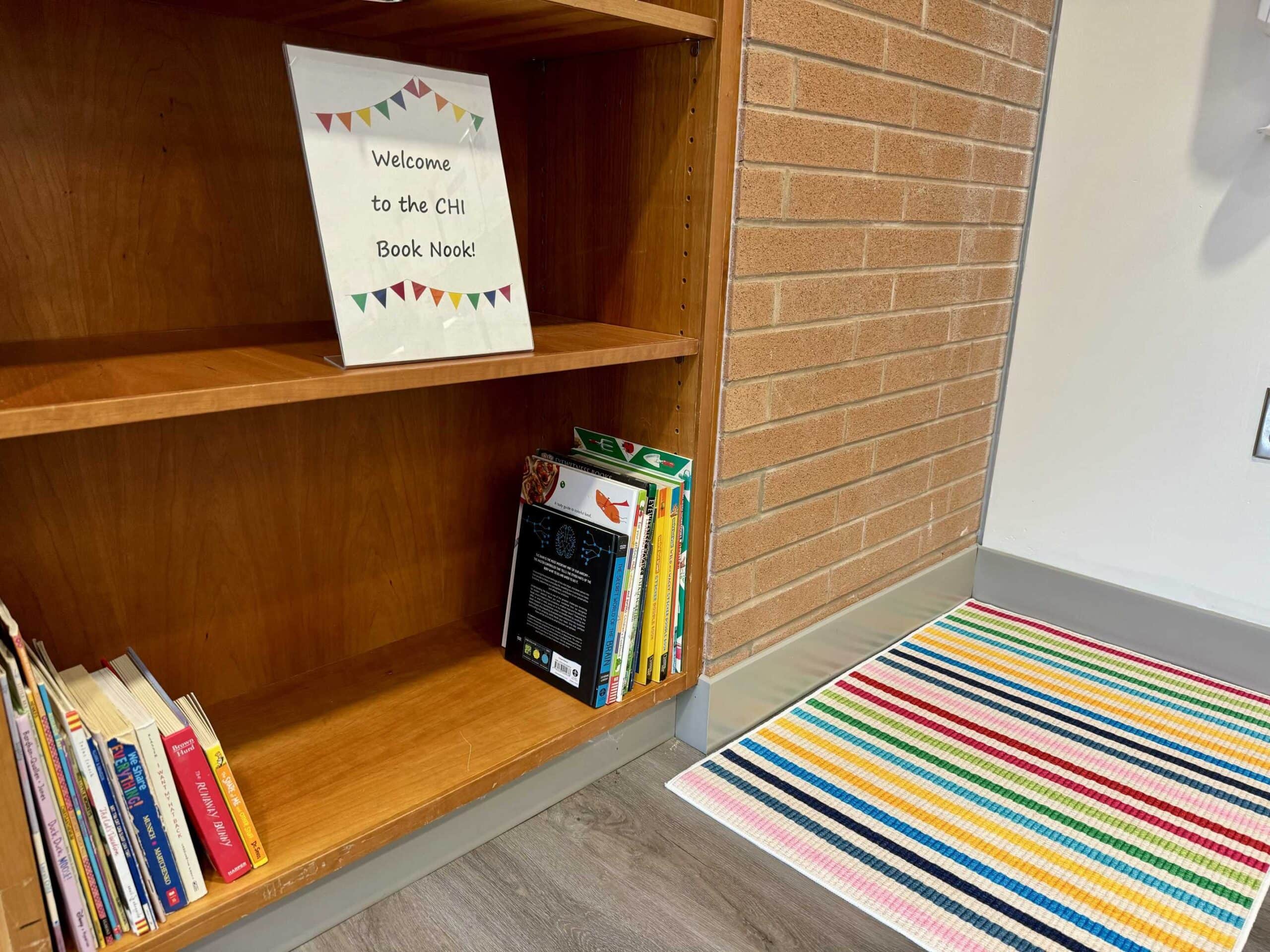 A cozy reading nook with a wooden shelf, books, a welcome sign, and a colorful striped rug on the floor.