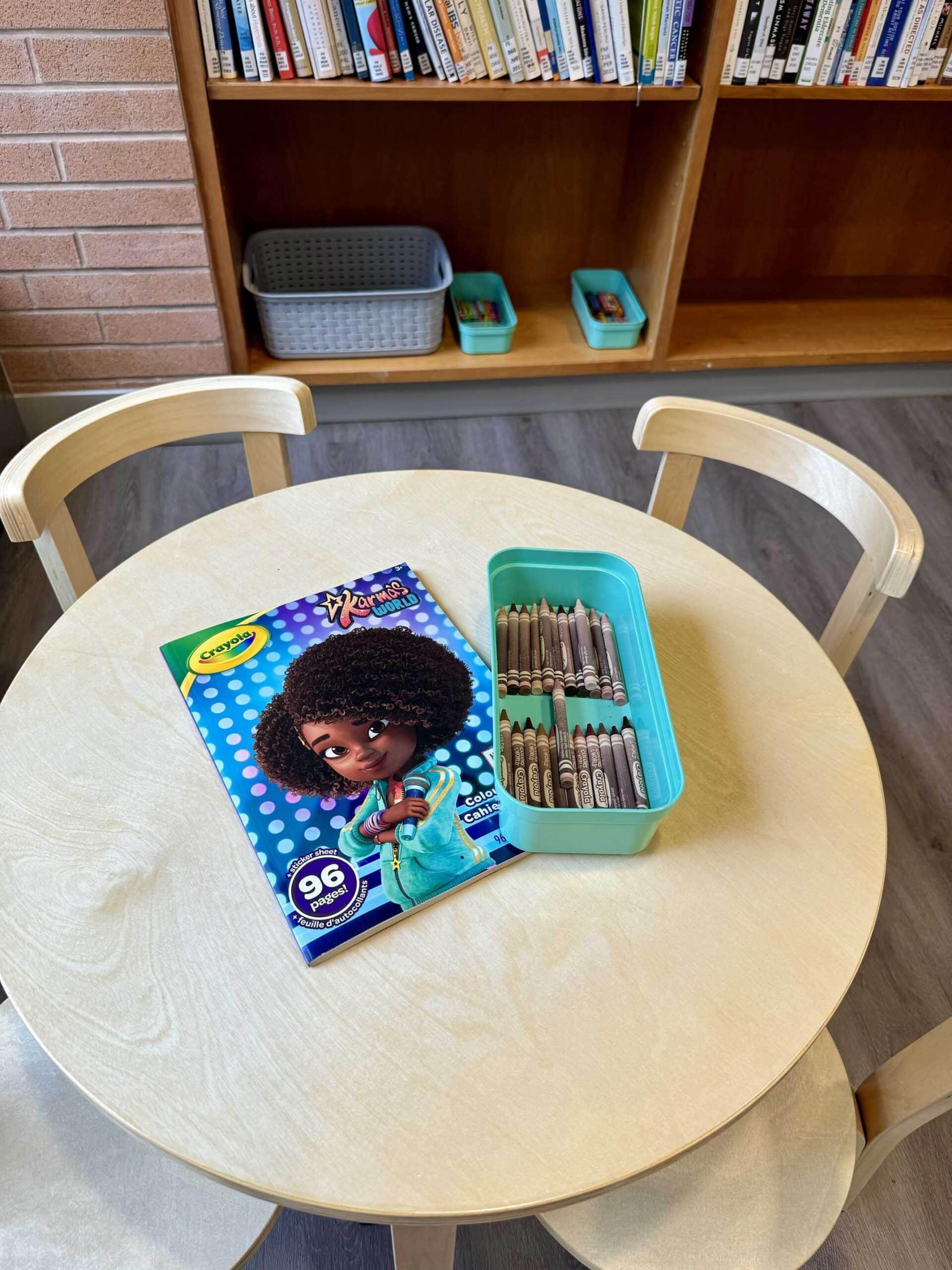 A round wooden table with two chairs, featuring a coloring book and a box of crayons. A bookshelf is in the background.