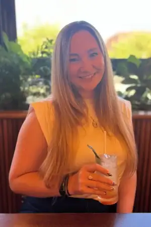 Long brown haired woman sat in front of a window. Wearing a white t-shirt and with a lemonade in front of her. Victoria Steele, Marketing Consultant