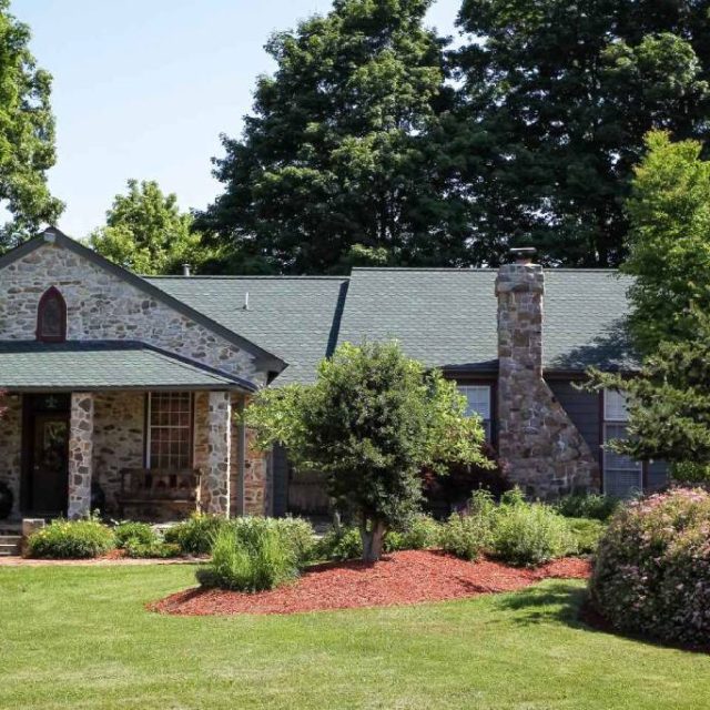 beautiful home with stone walls and new gray roof surrounded by lawn and trees