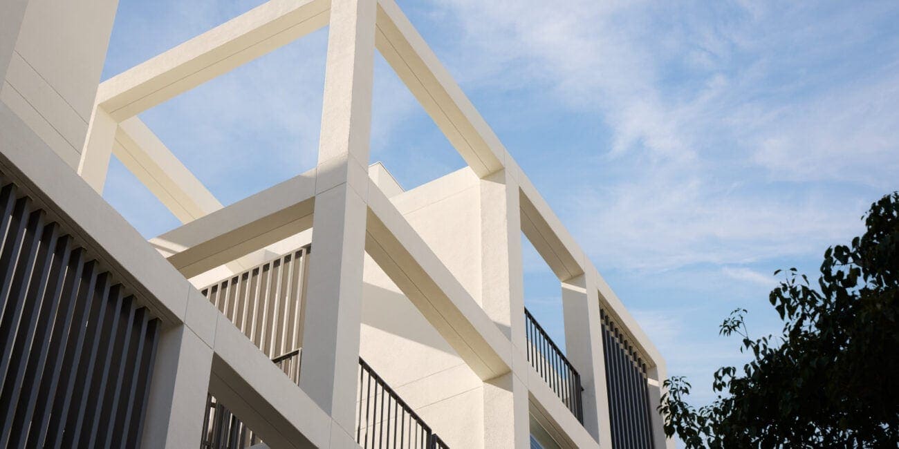 Modern white concrete building with large geometric open frames and black metal balcony railings under a blue sky.