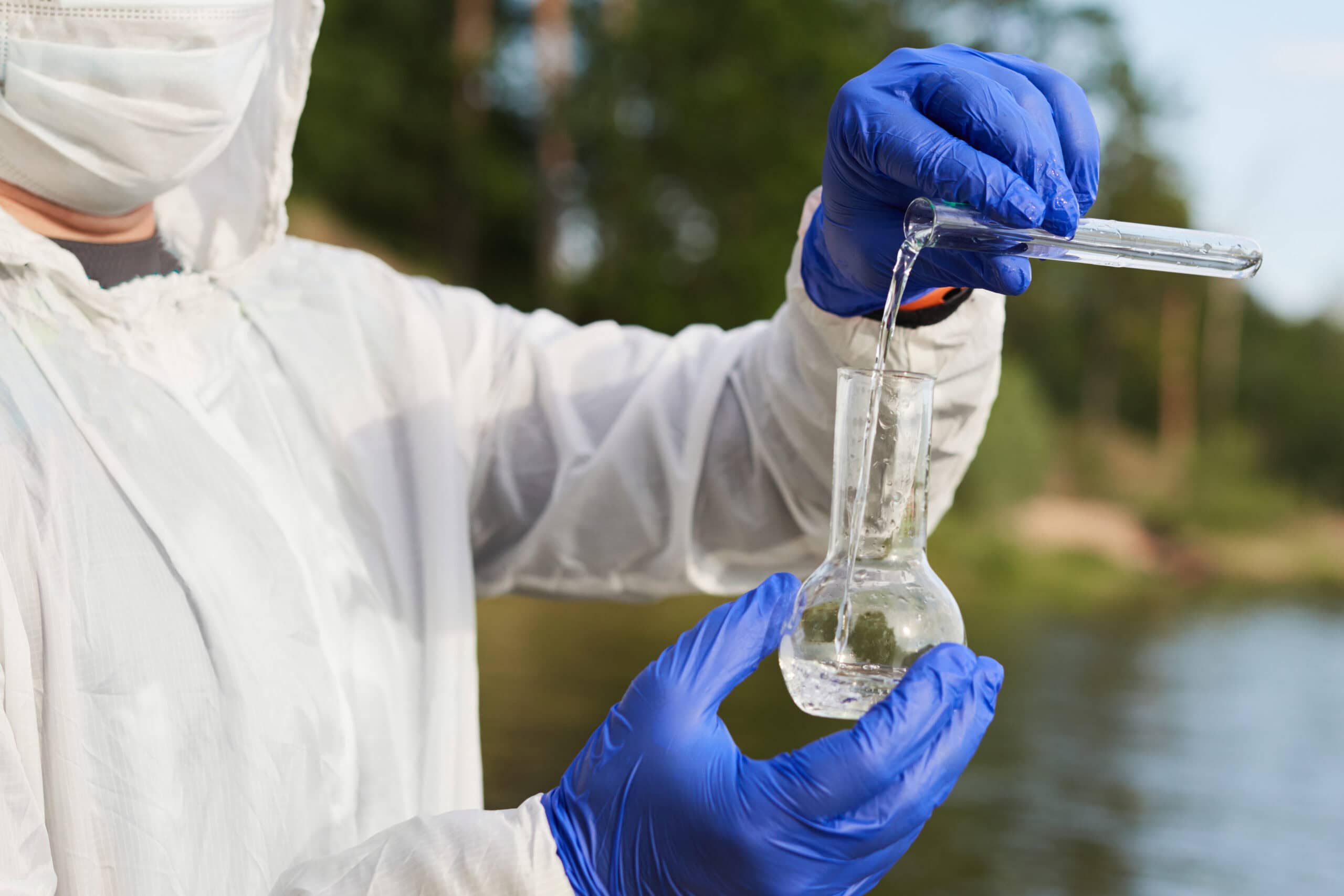 Water Purity Test. Hand in protective gloves holding a chemical flask or test tube with water. Lake in the background. Water Purity Test. Hand holding a chemical flask or test tube with water. Lake or river in the background.