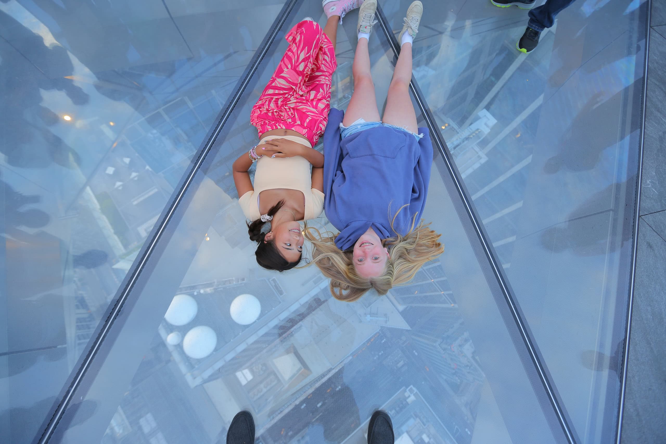 Two girls laying down on the glass floor of The Edge smile for a photo