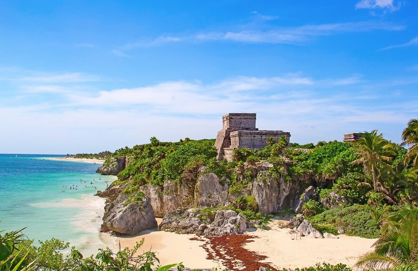 The Tulum cliffside temple overlooking a pink-tinted sandy beach with rocks, people swimming in the turquoise sea and a clear blue sky.