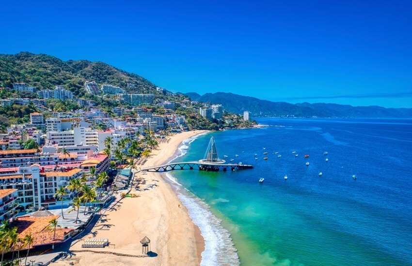 Puerto Vallarta beach with hotels and apartments along the shore and hillside, deep blue water, a clear blue sky and a few boats near the pier.
