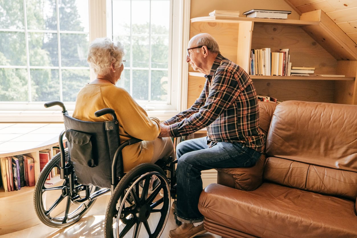 In a brightly lit room, a male caregiver sits on a leather couch while reaching out to touch a woman sitting in a wheelchair
