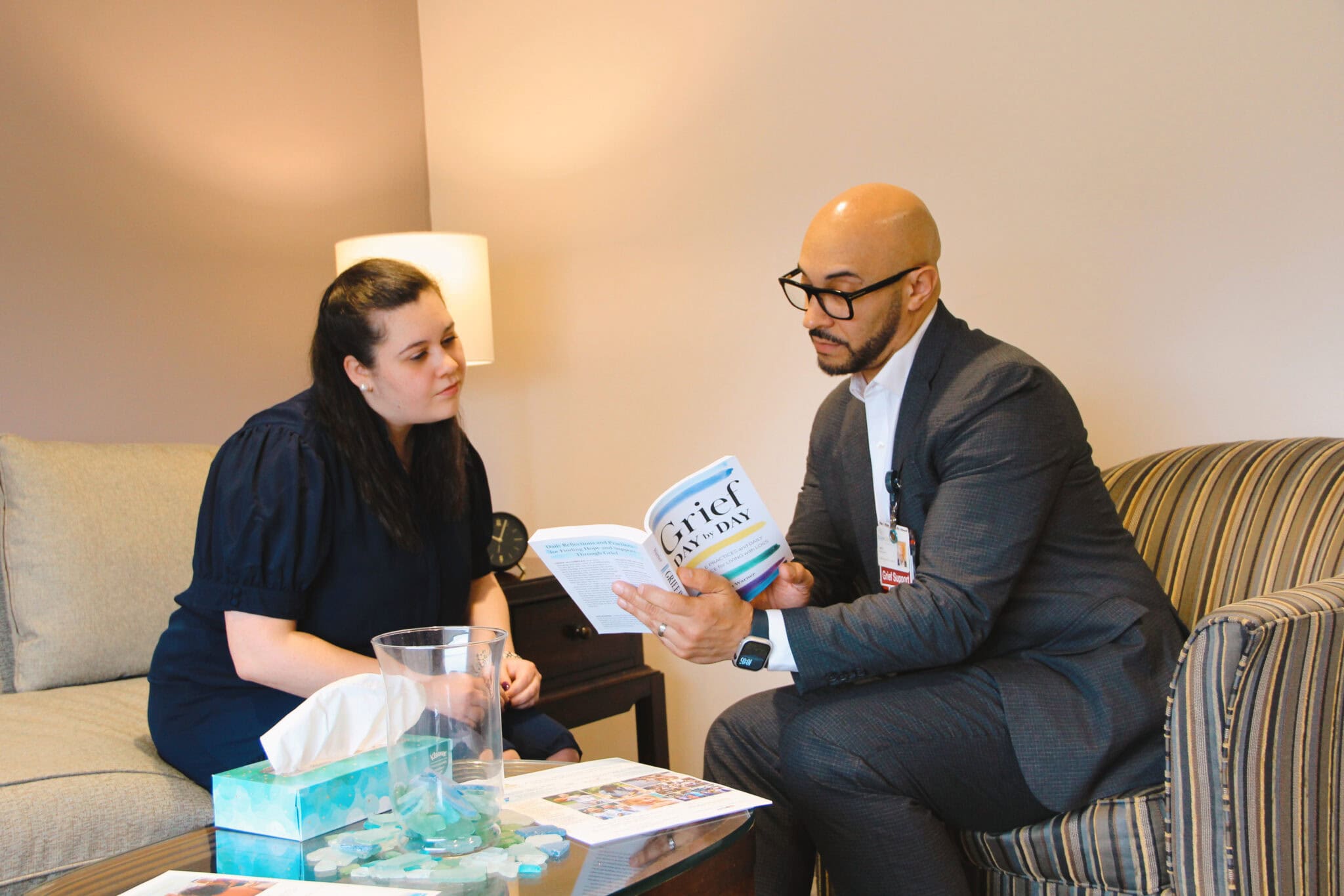Male grief counselor sits with a young woman, showing her a passage in a grief support book as they both look at it together.