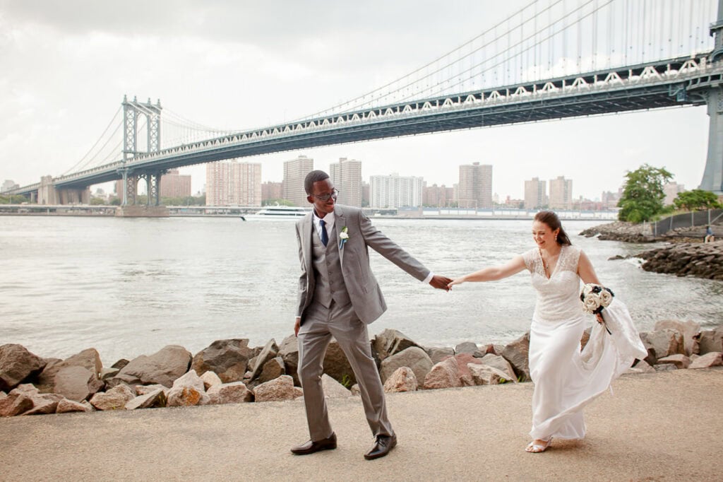 A bride and groom holding hands in front of the Manhattan Bridge during their wedding day.