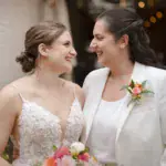 Two brides smiling at each other; one is wearing a white dress and the other a white suit. Both are holding a flower bouquet.