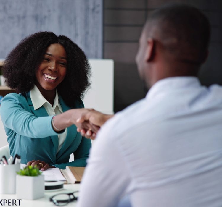 A woman and a man shake hands at a desk, symbolizing a professional meeting or interview setting.