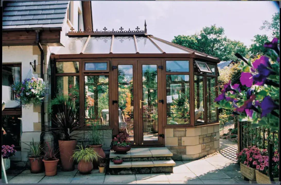 Brown Victorian conservatory with glazed roof, double doors and stone base surrounded by garden plants.