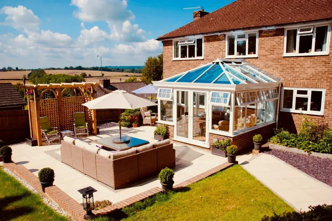 White conservatory with a blue-tinted glass roof, French doors and a paved garden patio with outdoor seating.