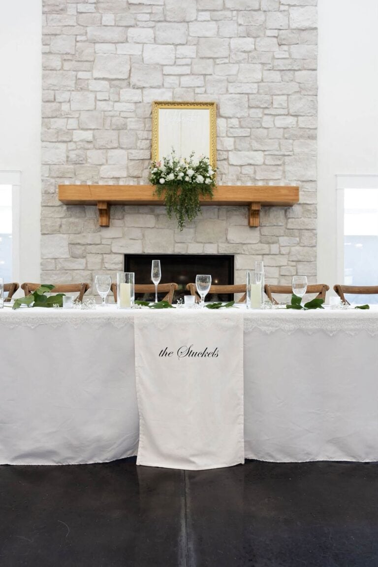 Head table with white linens and greenery in front of the stone fireplace in the Romaine Valley reception hall