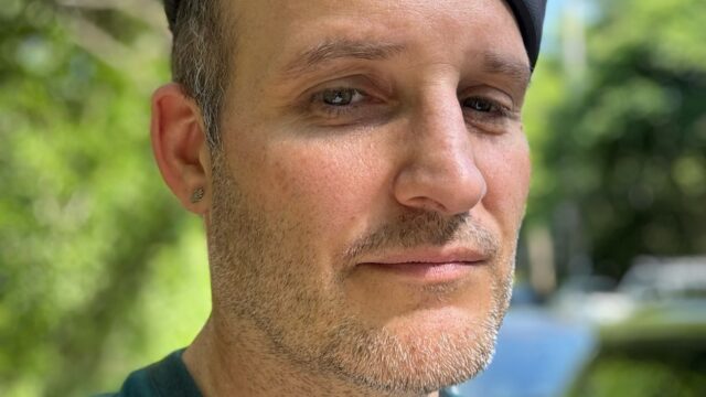Jess Conn-Potegal wearing a dark cap and teal shirt, gazing calmly at the camera in a sunlit outdoor setting with trees in the background.