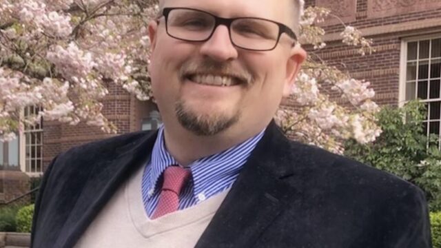 Joshua Fitzgerald smiling outdoors in front of a brick building and blooming cherry blossoms, wearing glasses, a navy blazer, a cream sweater vest, and a red tie.