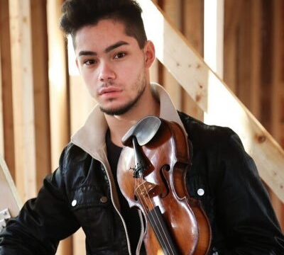 Violinist Royce Rich poses with a violin in a wooden interior, wearing a black jacket with a beige collar and looking directly at the camera.