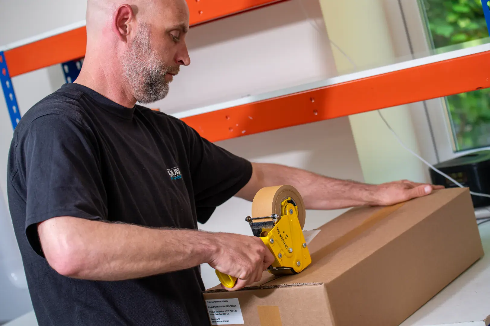 Worker sealing a long cardboard box with a tape dispenser in a packing area.