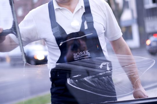 Auf dem Bild sieht man einen Mann von der Brust bis zur H&uuml;fte, der offenbar ein Fenster reinigt. Er tr&auml;gt ein wei&szlig;es Poloshirt und eine dunkle Sch&uuml;rze mit Tr&auml;gern. Auf der Sch&uuml;rze steht &bdquo;BOREK&ldquo; sowie kleinere Schrift darunter.
Der Mann h&auml;lt ein Werkzeug in der Hand, vermutlich einen Fensterabzieher oder ein Reinigungsger&auml;t. Auf der Glasscheibe sind deutliche Wischspuren und Seifenreste zu sehen. Im Hintergrund erkennt man unscharf eine Stra&szlig;e mit Autos und Geb&auml;uden, was darauf hindeutet, dass die Aufnahme drau&szlig;en oder an einer gro&szlig;en Fensterfront entstanden ist.