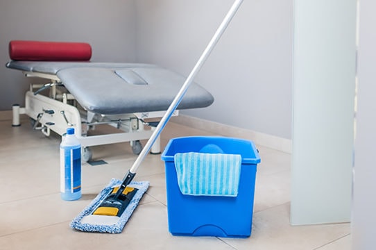 Auf dem Bild sieht man einen Reinigungsbereich in einem medizinischen oder therapeutischen Raum. Im Hintergrund steht eine Behandlungsliege mit grauer Polsterung und einer roten Kopfst&uuml;tze.
Im Vordergrund befinden sich Reinigungsutensilien: ein blauer Putzeimer mit einem Tuch &uuml;ber dem Rand, ein Wischmopp mit Stiel sowie eine Flasche Reinigungsmittel. Der Boden ist hell und wirkt sauber.
Die Szene vermittelt den Eindruck, dass der Raum gerade gereinigt wird oder f&uuml;r die n&auml;chste Nutzung vorbereitet wird, zum Beispiel in einer Arztpraxis oder einem Behandlungsraum.