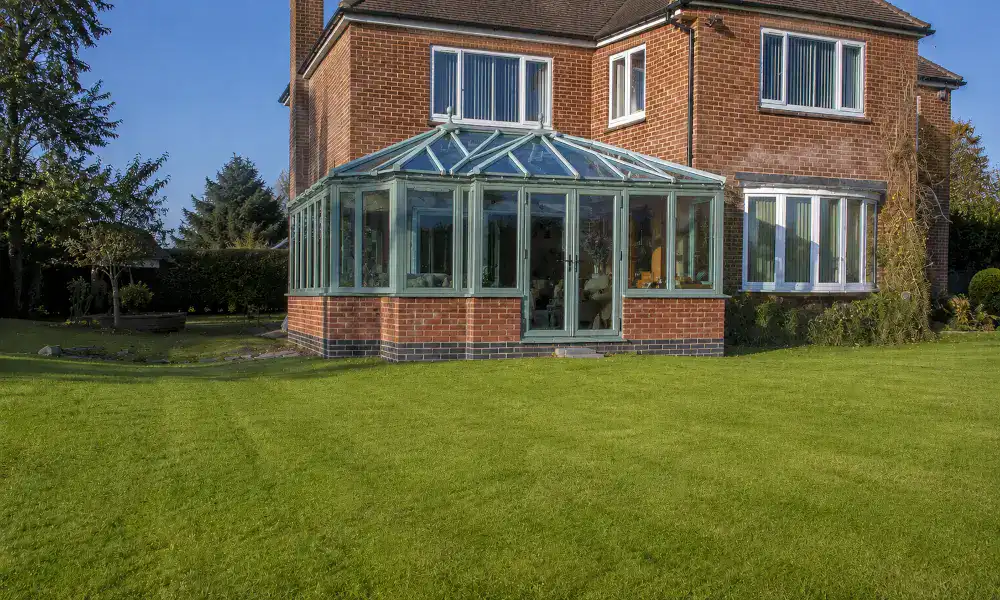 High-angle aerial drone view of a bespoke, Chartwell green uPVC conservatory built in an Edwardian style, featuring a multi-faceted glass roof and double doors, attached to a red brick house with a green lawn.