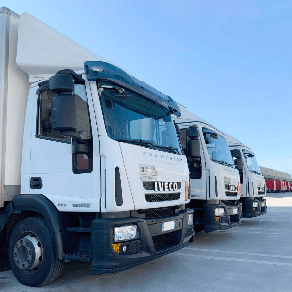 Tre camion Iveco bianchi appartenenti alla M.C. AUTOTRASPORTI parcheggiati in fila, con un cielo blu sullo sfondo.