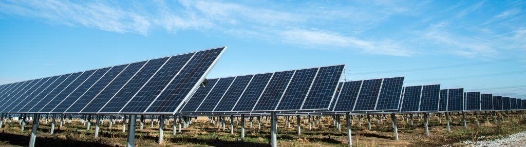 A ground-level, horizontal view of many rows of dark blue solar panels angled toward a bright blue sky with wispy clouds.