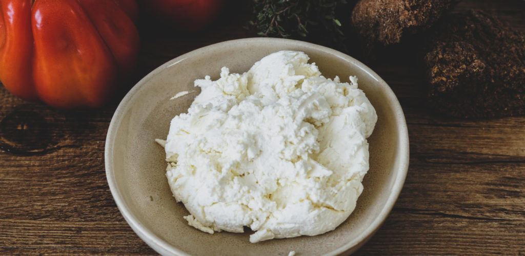 Frischkäse im Schälchen auf einem Holztisch.Tomaten und griechisches Zwieback im Hintergrund