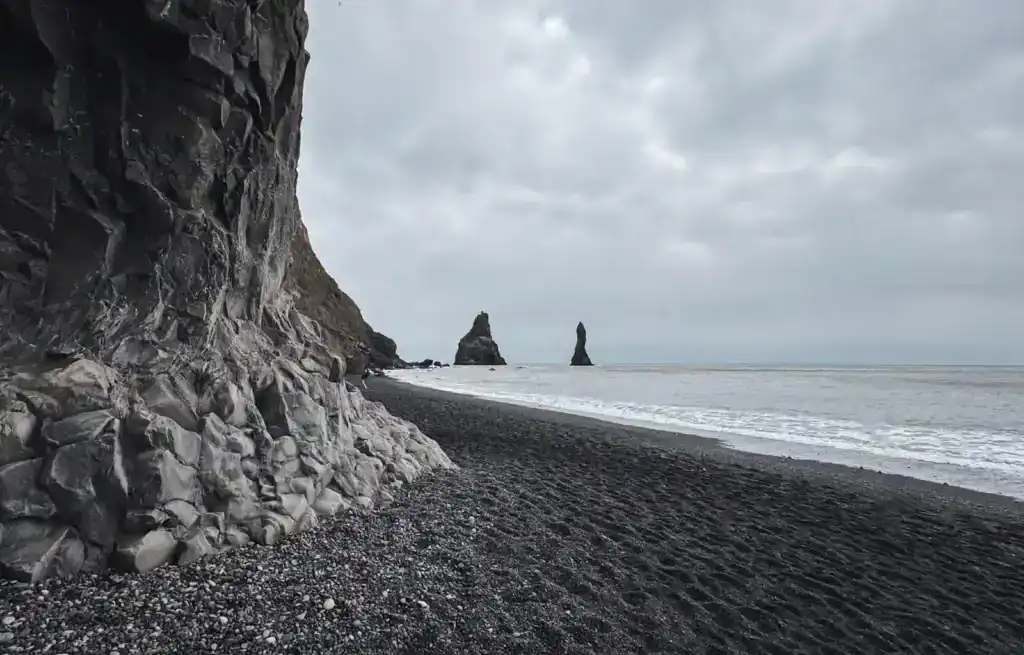 Dramatic basalt cliffs rise along the edge of Reynisfjara, Iceland’s famous black sand beach. The dark volcanic shoreline stretches toward the ocean, where powerful waves crash against the shore. In the distance, the Reynisdrangar sea stacks emerge from the misty waters, standing tall against the overcast sky. The moody atmosphere enhances the beach’s rugged and otherworldly beauty.