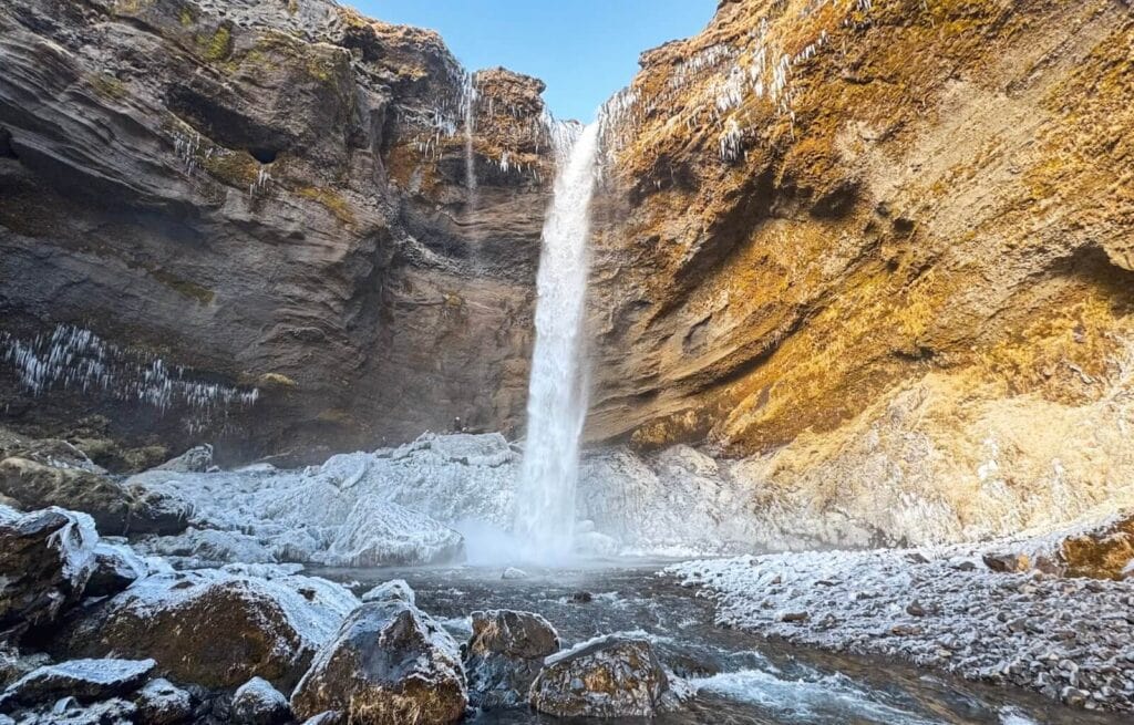 Kvernufoss Waterfall cascading down a steep cliff into a rocky pool below, surrounded by rugged cliffs and clear blue sky.