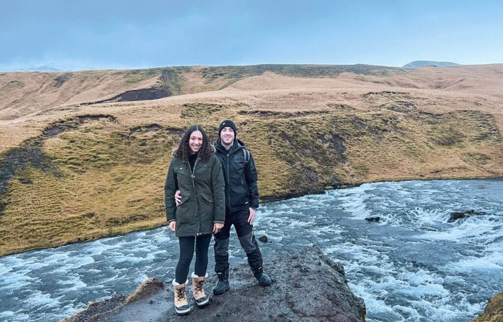 Two people standing on rocks beside a flowing river with grassy cliffs in the background.