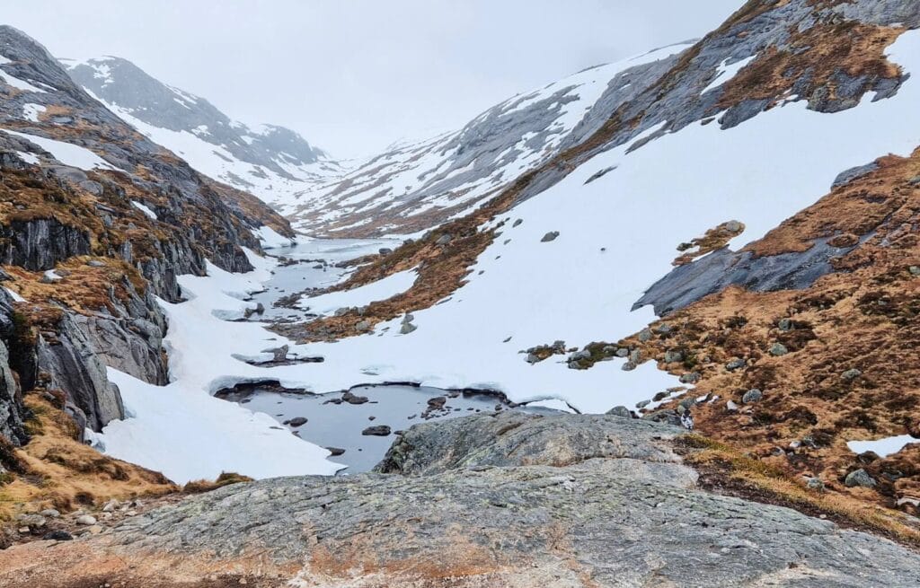 Snow-covered mountain valley with rocky slopes and a small stream flowing through the centre.