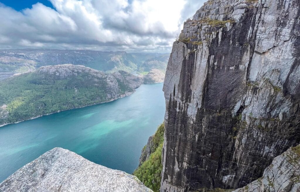 Cliffside view from a dramatic vantage point in Norway, showcasing a deep fjord with turquoise-blue waters framed by towering rock faces and lush green hills. Low-hanging clouds drift above the mountainous terrain, capturing the raw beauty highlighted in our Norway travel guide. Ideal for hiking, sightseeing, and nature photography.