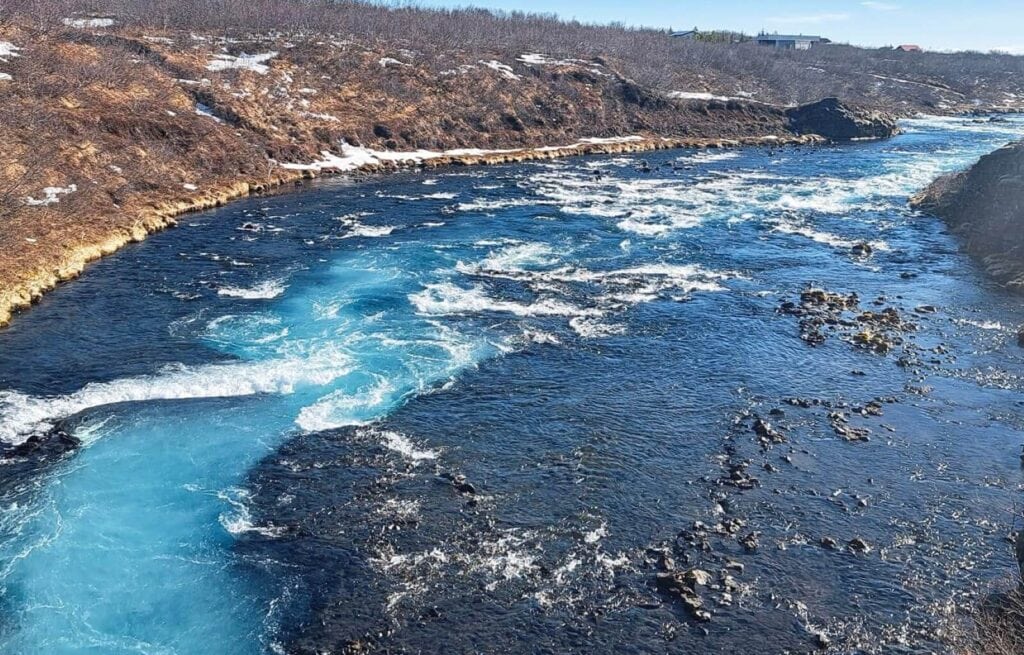 Wide view of Brúarfoss River with blue water and rocky banks, snow patches on the hillside in the background.