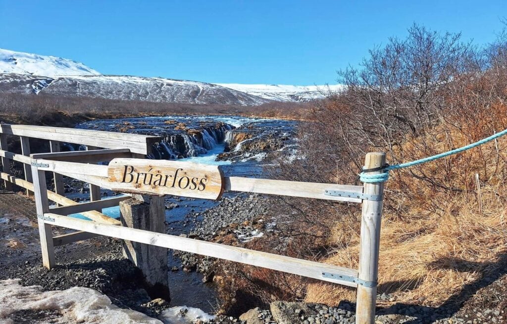 View of Brúarfoss Waterfall with a wooden fence and a sign in front, snow-capped mountains in the background under a clear, 蓝.