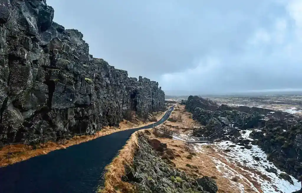 Pathway through rocky cliffs in Thingvellir National Park with snow patches and cloudy sky.