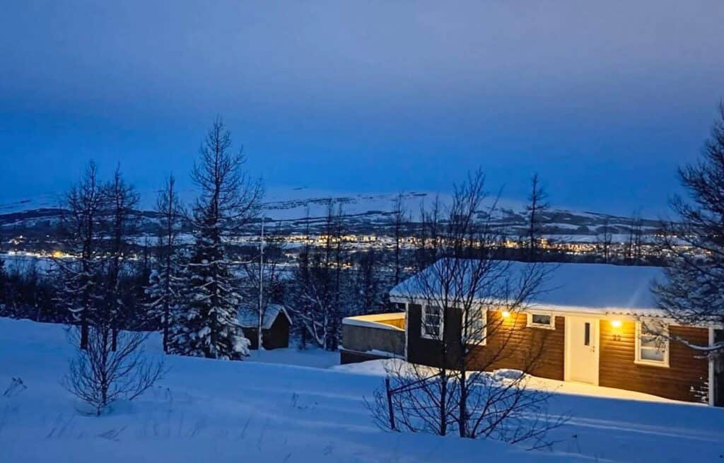 Snow-covered landscape with a cosy wooden house illuminated at night, surrounded by leafless trees and distant hills.