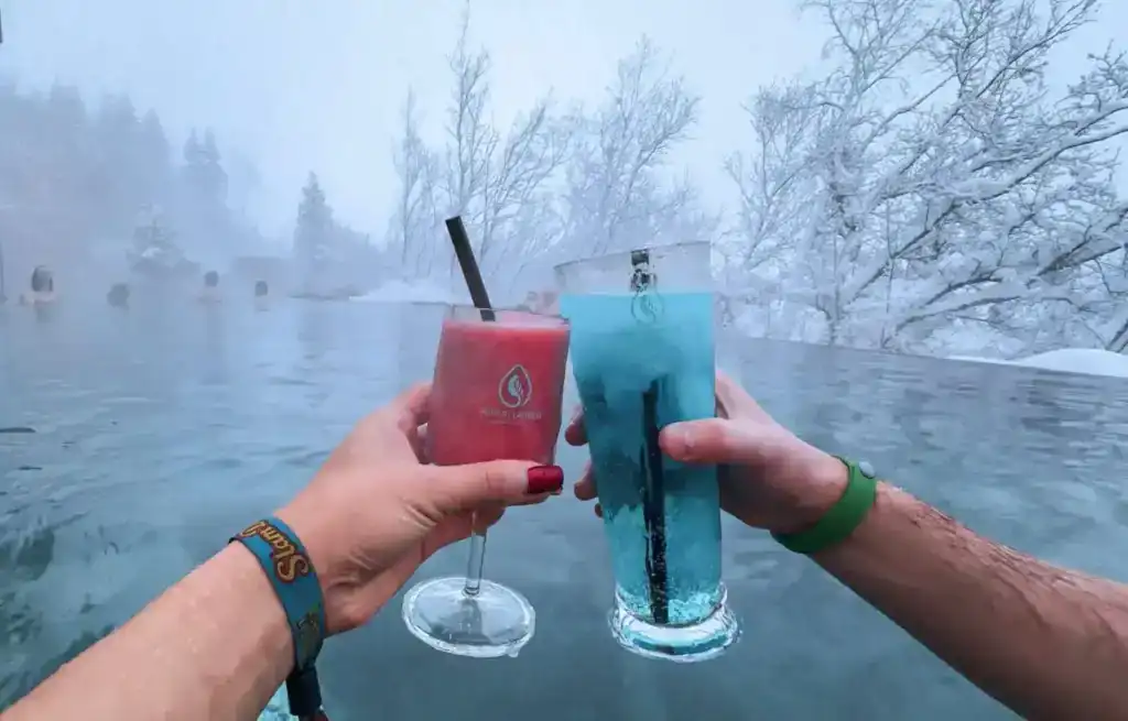 Two hands holding colourful drinks in hot spring with snowy trees in background.