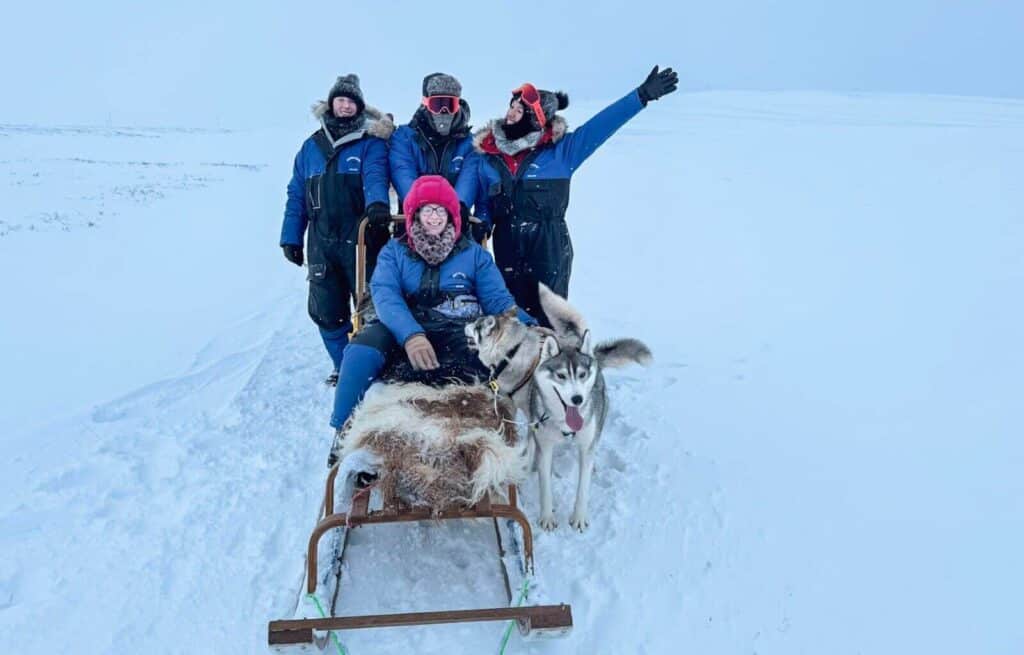 Group of four people in winter gear riding a dog sled across snow.