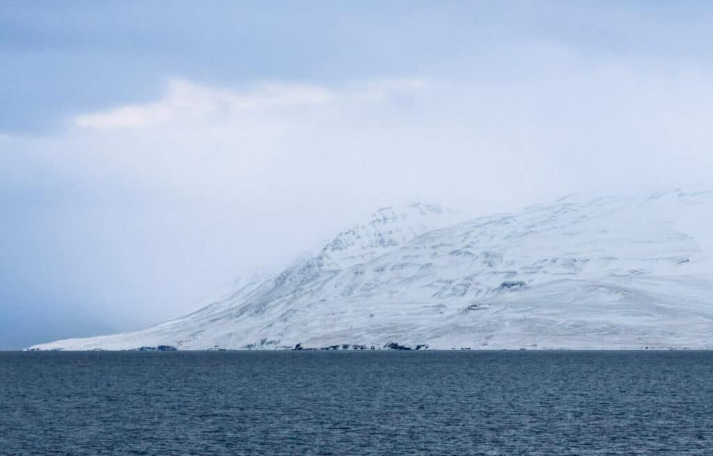 Snow-covered mountain slopes meet the calm sea under a cloudy sky in Akureyri.