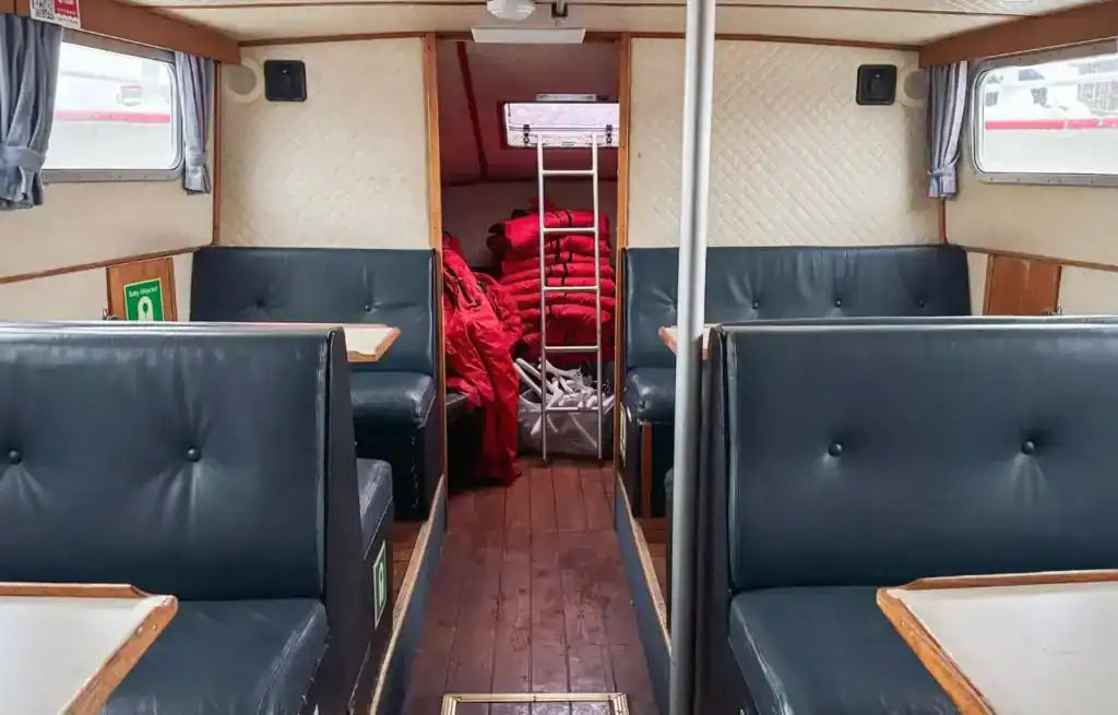 Inside a vintage train carriage with black leather seats and wooden tables, showing a corridor with red blankets and luggage.