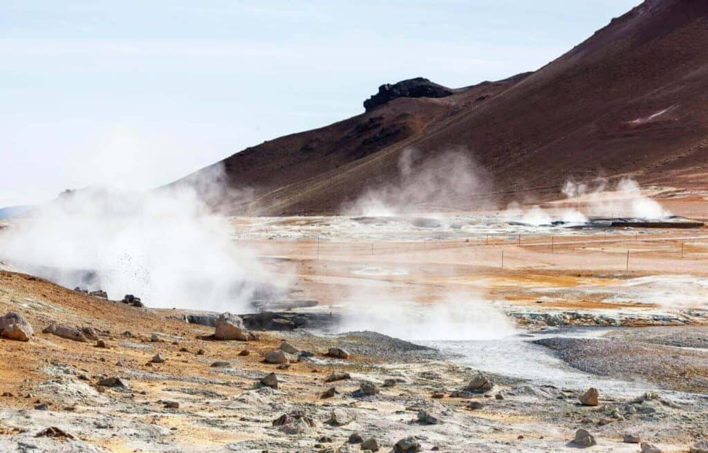 Geothermal area with steam rising from the ground and rugged volcanic landscape in the background.