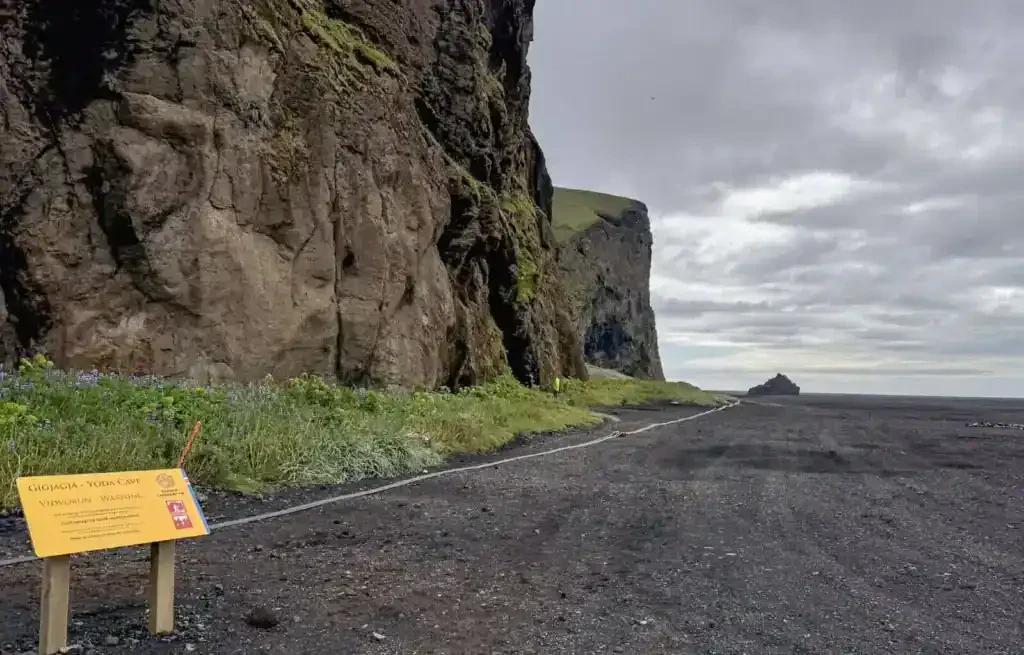View of a black sand beach with a cliff on the left and cloudy sky above.
